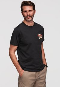 Homme en t-shirt noir avec un dessin de crâne et de roses sur la poitrine, pantalon beige, debout les mains dans les poches sur un fond clair uni.