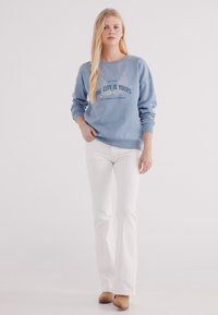 Felpa azzurro chiaro con la scritta "LA CITTÀ È TUA" stampata in blu scuro, abbinata a pantaloni bianchi a campana e scarpe beige. Tessuto dalla texture liscia.
