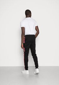 Svarta joggingsbyxor med röda sidostreck och fickor, tillverkade av ett mjukt material, kombinerat med en vit t-shirt och vita sneakers på en neutral bakgrund.