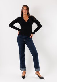 Top lungo a maniche ribbed nere con scollatura a V e dettagli in metallo argentato, abbinato a jeans blu a gamba dritta con risvolti e scarpe nere a punta.
