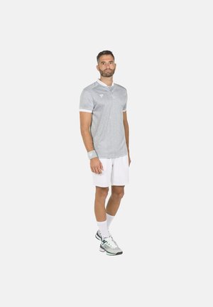 Hombre de pie con camiseta polo gris, pantalones cortos blancos, pulsera, calcetines blancos y zapatillas grises sobre un fondo blanco liso.