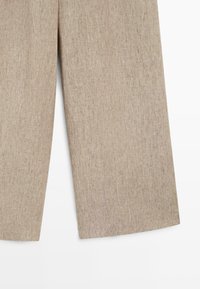 Beige linnen broek met wijd uitlopende pijpen, weergegeven vanaf de knie naar beneden op een witte achtergrond.