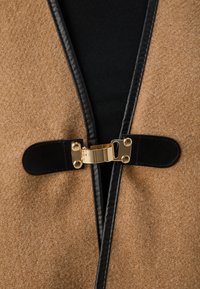 Tan vævestof i uldblanding med sort læderbesætning, der har en skinnende guldkrog og en struktureret overflade, der viser et moderne design.
