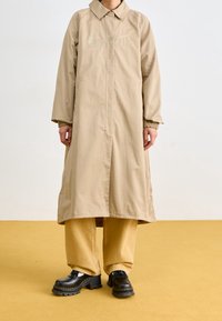 Béžový dlouhý kabát s límcem, zapínáním na zip a bočními kapsami; má kovové logo; doplněný širokými khaki kalhotami a černými platformními botami.