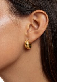 Goldfarbene Creolen mit einer glatten, reflektierenden Oberfläche und einem geschwungenen, nierenförmigen Design, die am Ohr getragen werden.