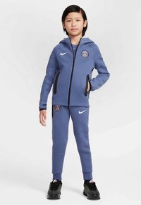 Nike Paris Saint-Germain træningssæt i blå, med en hættetrøje med lynlås og logo, bukser med sidelommer og sorte sneakers. Glat tekstur.