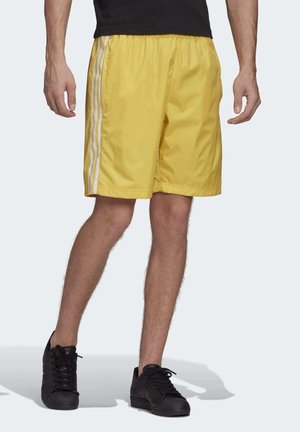 Homme portant un short de sport jaune avec des bandes blanches sur les côtés et des baskets noires, debout les jambes écartées sur un fond clair.