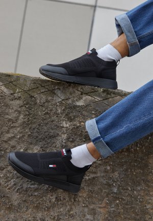 Piernas con vaqueros azules remangados, calcetines blancos y zapatillas negras de punto sin cordones descansando sobre una superficie de hormigón texturizado.