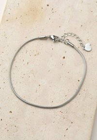 Pulsera de metal plateado con un diseño suave y flexible, que cuenta con un cierre de langosta y una cadena de extensión con un pequeño charm redondo.