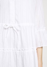 Robe blanche à rayures verticales et points texturés, avec ceinture à la taille et manches cloche trois-quarts.