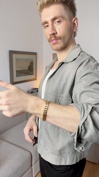 Metalni ručni sat s pravokutnim dizajnom zlatne boje. Model nosi svijetlosivu jaknu preko bijele košulje, u opuštenoj pozi.