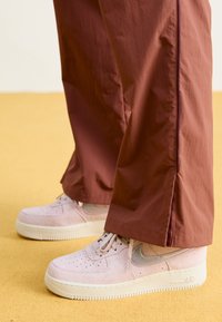 Sneakers in suede rosa chiaro con suola bianca e dettagli argentati, abbinati a pantaloni impermeabili borgogna a taglio ampio. Sfondo giallo.