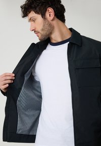 Hombre con camiseta blanca muestra chaqueta negra con forro de cuadros vichy, mirando hacia su izquierda.