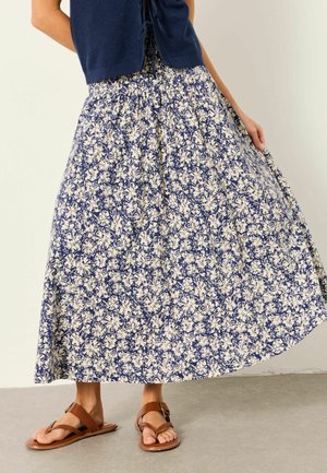 Frau trägt einen marineblauen Midirock mit Blumenmuster und gerafftem Bund, ein ärmelloses navyfarbenes Oberteil und braune Ledersandalen und steht auf einem grauen Boden.