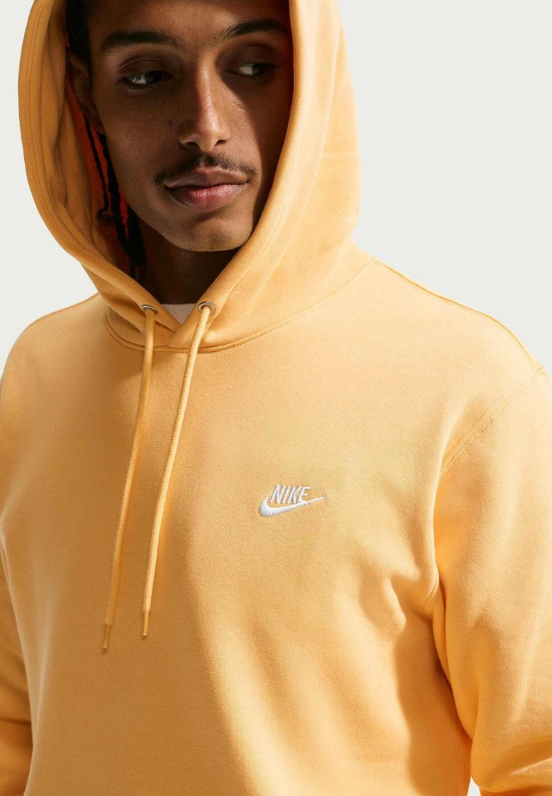 Junger Mann mit Schnurrbart schaut nach unten, trägt einen gelben Nike-Hoodie mit aufgesetzter Kapuze und sichtbaren Kordeln vor einem einfarbigen Hintergrund.