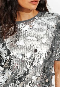 Haut à sequins argentés avec une coupe décontractée, manches courtes et un col rond bordé de gris. Le tissu est réfléchissant et texturé.