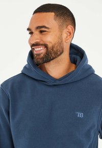 Marineblauwe fleece hoodie met een kangoeroe-pocket, verstelbare capuchon en een klein geborduurd logo op de borst. Zachte textuur en casual design.