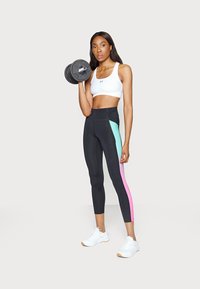 Zwarte hoge taille leggings met mintgroene en roze zijpanelen, gecombineerd met een witte sport-bh. Gewichte dumbbells in één hand vastgehouden.