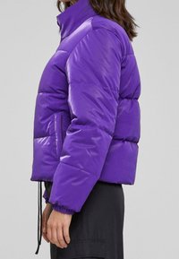 Fioletowa pikowana kurtka z wysokim kołnierzem, elastycznymi mankietami i regulowanym sznurkiem u dołu. Gładka, błyszcząca powierzchnia i krótki krój.