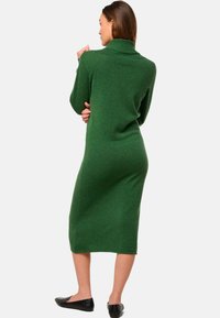 Grünes gestricktes Sweatkleid mit hohem geripptem Kragen, langen Ärmeln und mittellanger Länge. Weiche Textur, figurbetonte Silhouette und seitliche Schlitze.