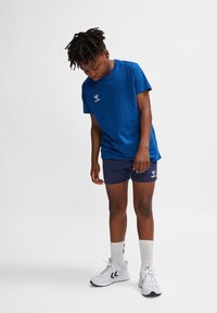 Blå bomulls-T-shirt, kortärmad, rund halsringning; ihop med marinblå shorts. Bärs med vita sneakers och ankellånga strumpor.