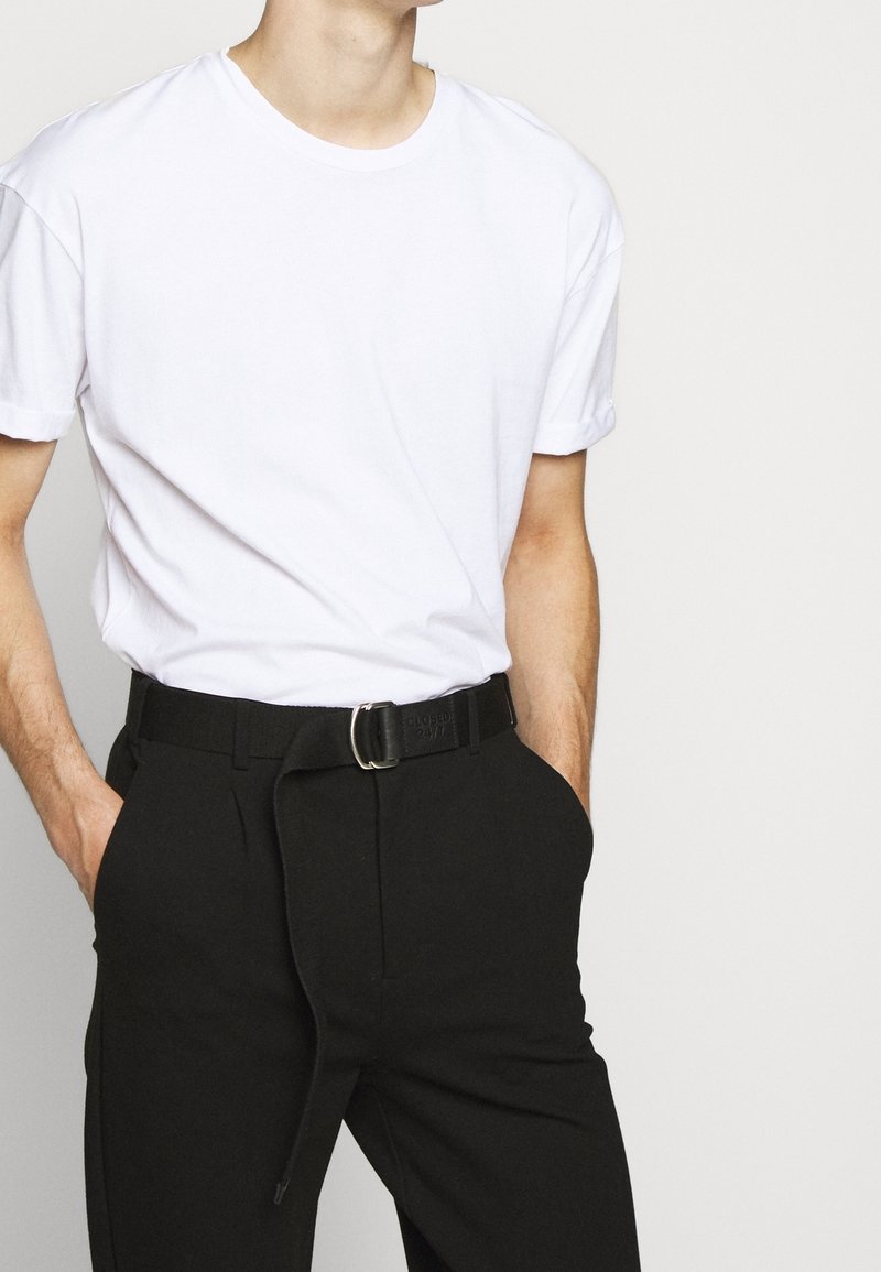 T-shirt en coton blanc avec manches retroussées, associé à un pantalon noir doté d'une ceinture avec une boucle argentée et d'un détail en tissu à la taille.