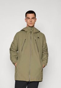 Chaqueta impermeable verde oliva con capucha, que cuenta con dos bolsillos frontales con cremallera y cierre de cremallera. Incluye una sutil marca en el pecho.