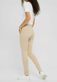 Kobieta z długimi kręconymi włosami, ubrana w białą koszulkę, beżowe slim-fit spodnie, białe sneakersy, stoi z rękami w kieszeniach.