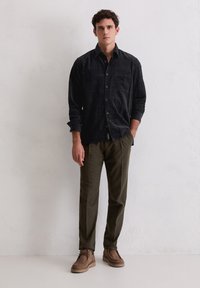 Czarna koszula w kratę z długim rękawem i kieszenią z przodu, zestawiona z oliwkowozielonymi spodniami i brązowymi zamszowymi butami na jednolitym tle.