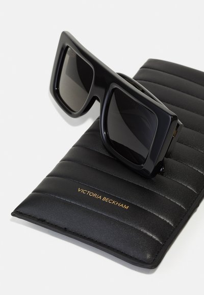 Black oversized square sunglasses resting on a black padded case with gold "Victoria Beckham" lettering.