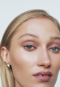 Gros plan d'un visage avec une peau claire et des cheveux blonds clairs, présentant des sourcils bien définis, un fard à paupières rose subtil et des boucles d'oreilles en argent.