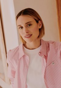 Νεαρή γυναίκα με κοντά ξανθά μαλλιά, ντυμένη με ροζ ριγέ πουκάμισο πάνω από λευκή μπλούζα, στέκεται σε εσωτερικό χώρο με ουδέτερο φόντο.