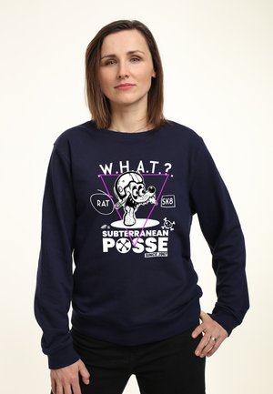 Frau trägt dunkelblauen Sweatshirt mit Cartoon-Ratte im Helm und Text „W.H.A.T? Subterranean Posse seit 1967“, steht mit Hand in der Hüfte.