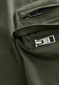 Tissu vert olive présentant un gros plan de fermetures éclair, de ferrures métalliques et une texture douce. Les coutures sont visibles près des poches.