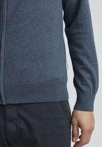 Mørkeblå strikket cardigan med glidelås, ribbede mansjetter og nederkant, med et lite merke på siden; laget av mykt, teksturert materiale.