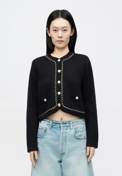 Veste noire courte avec détails en métal doré, dotée d'un col rond, bordée d'une chaîne et de deux poches avant. Texture lisse et coupe ajustée.