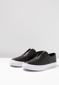 Une paire de baskets noires à enfiler avec un dessus en cuir texturé et de épaisses semelles en caoutchouc blanc, sur une surface blanche avec un fond uni.