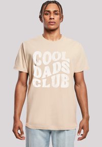 Camiseta de algodón beige con gran texto blanco desgastado "CLUB DE PADRES GENIALES" en la parte frontal. Mangas cortas y cuello redondo.