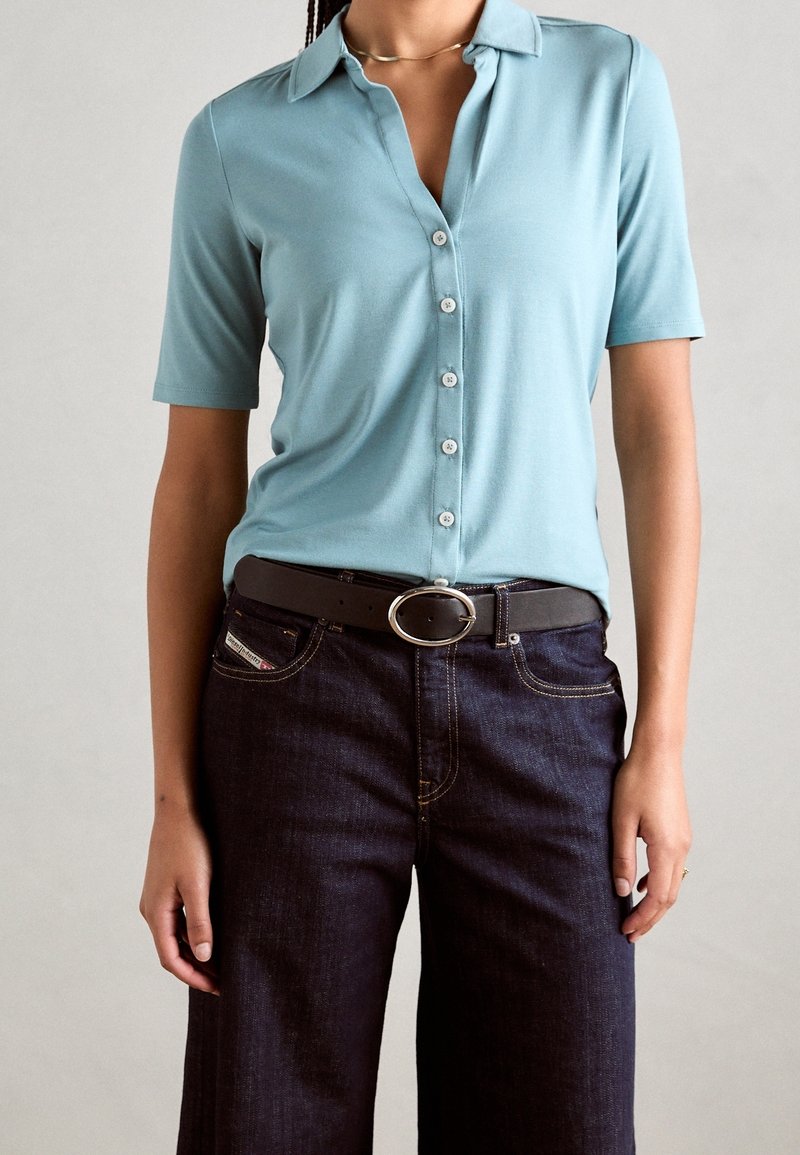 Polo shirt azzurro chiaro a maniche corte con colletto e pattina a bottoni, abbinato a jeans in denim scuro e una cintura marrone con fibbia ovale in argento.