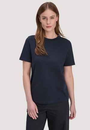 Junge Frau mit glattem hellbraunem Haar, die ein schlichtes dunkelblaues T-Shirt mit kurzen Ärmeln und schwarze Hosen trägt, steht vor einem hellen Hintergrund.