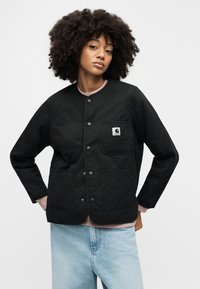 Giacca nera trapuntata con bottoni a pressione, dotata di una tasca sul petto e due tasche frontali. Indossata sopra una maglia rosa con scollo tondo e jeans azzurri chiari.