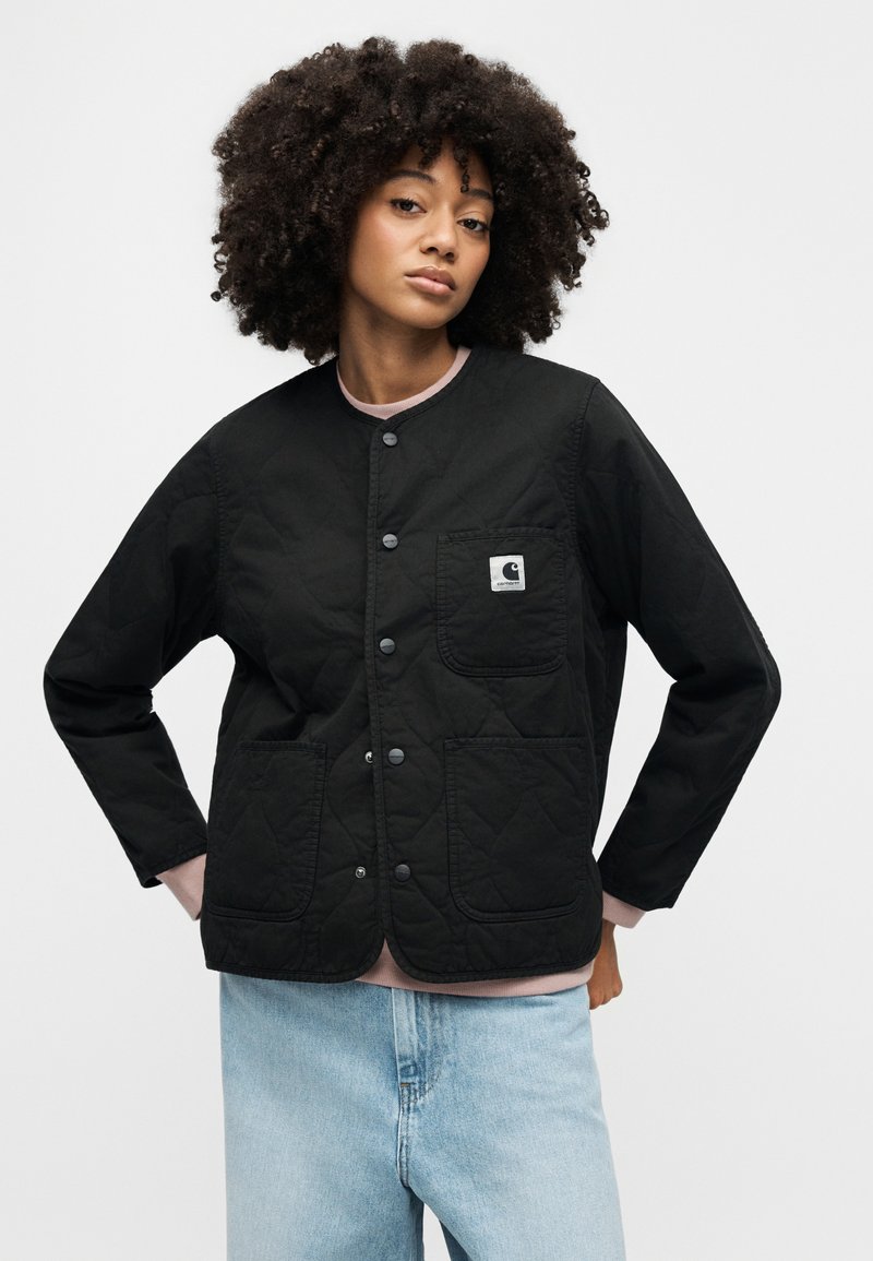 Zwarte doorgestikte jas met drukknopen, voorzien van een borstzak en twee voorzakken. Gedragen over een roze T-shirt met ronde hals en lichtblauwe spijkerbroek.