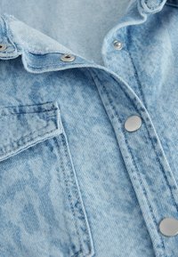 Tissu en denim bleu clair avec boutons-pression en métal et une poche à rabat, montrant des coutures détaillées et une texture.