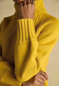 Person, die einen strukturierten gelben Strickpullover trägt, mit verschränkten Armen und einer Hand mit goldenen Ringen in der Nähe des Gesichts und der Taille.