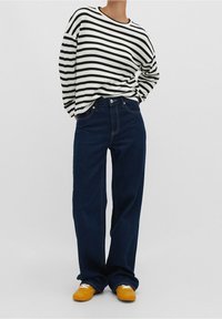Pull rayé noir et blanc, coupe décontractée, porté avec un jean large bleu foncé et des chaussures jaunes, avec des accents blancs sur les chaussures.
