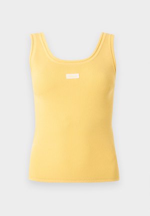 Γυναίκα που φοράει κίτρινη αμάνικη μπλούζα, καφέ παντελόνι, μαύρα μοκασίνια και κρατάει μια δερμάτινη μωβ τσάντα ώμου, στέκεται μπροστά σε απλό φόντο.