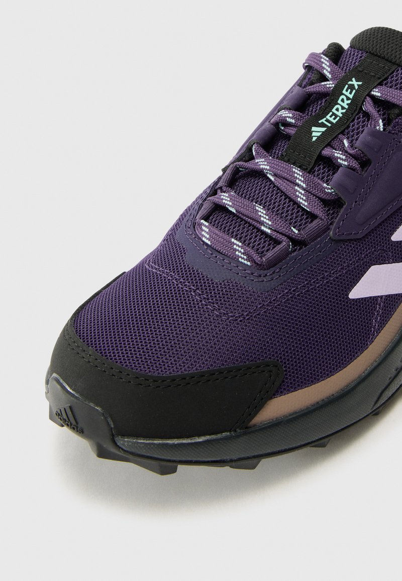 Zapatilla morada y negra con parte superior de malla, puntera de goma y suela texturizada. Presenta cordones reflectantes y una marca en la lengüeta.