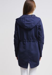 Veste à capuche bleu marine avec taille cintrée, détail élastique et dos légèrement plus long, réalisée en tissu lisse et résistant à l'eau.