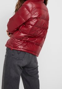 Rød dunjakke med blank finish, quiltet design og høj krave. Parret med mørkegrå jeans, der fremhæver en afslappet pasform.