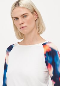 Kobieta z krótkimi blond włosami, ubrana w białą bluzkę z długimi rękawami w wielokolorowy, niebieski, czerwony i pomarańczowy wzór, patrząca na wprost na białym tle.